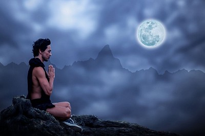 Bir kişinin meditasyon pozisyonunda ellerini göğsünde birleştirdiği silueti, ay ışığının aydınlattığı sakin bir dağ manzarası önünde. Gece gökyüzü ve bulutlar arka planda huzurlu bir atmosfer yaratıyor. Spiritüel ve doğa temalı, mavi-gri tonlarda bir görsel.